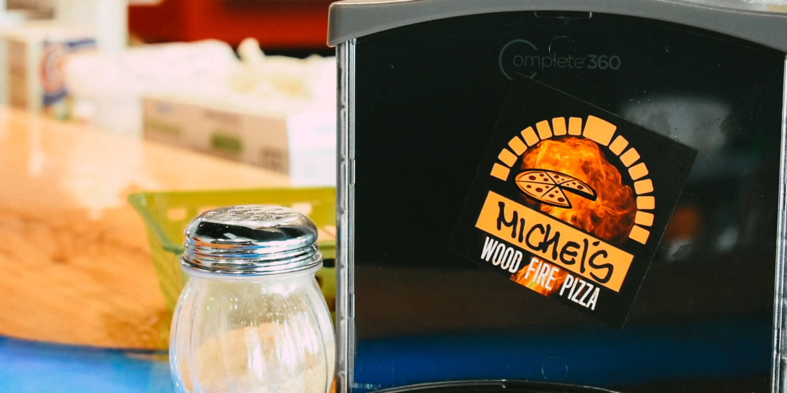 Logo for Michel's Wood Fire Pizza with a cartoon pizza and a flame is stuck to the side of a napkin dispenser, which is next to a shaker of parmesan cheese